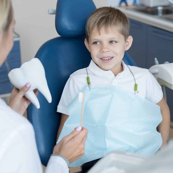 Portrait of smiling  little boy sitting in dental chair listening to female dentist holding toothbrush and tooth model explaining oral hygiene rules